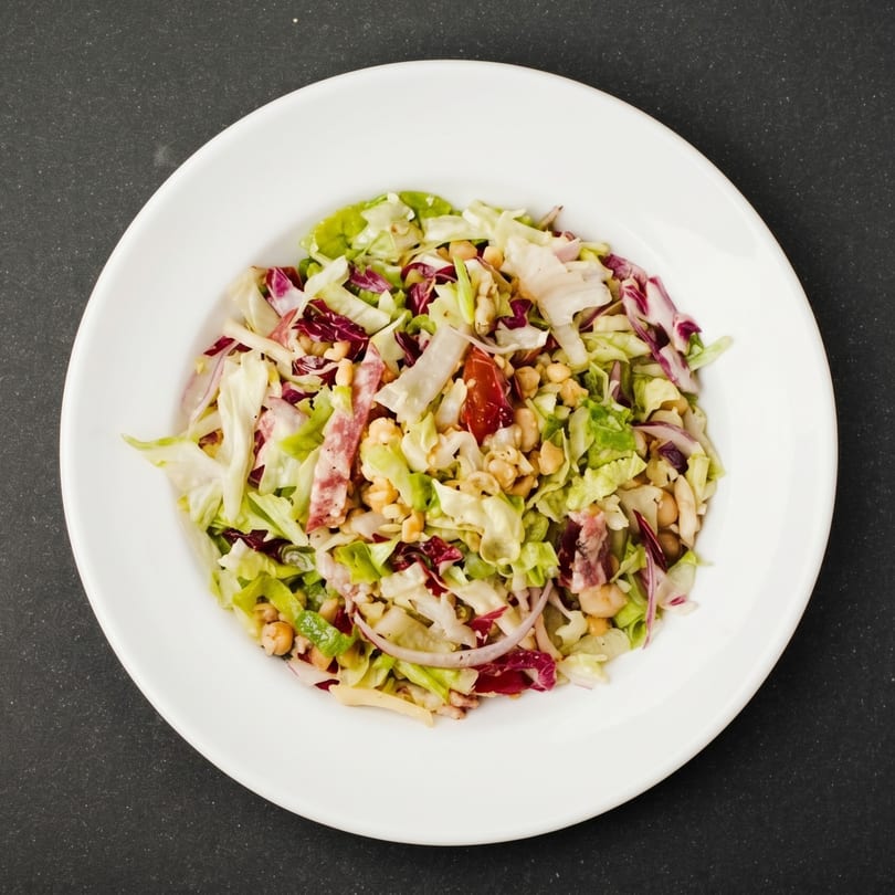 Overhead view of a vibrant salad with mixed greens, red cabbage, chickpeas, and salami on a white plate.