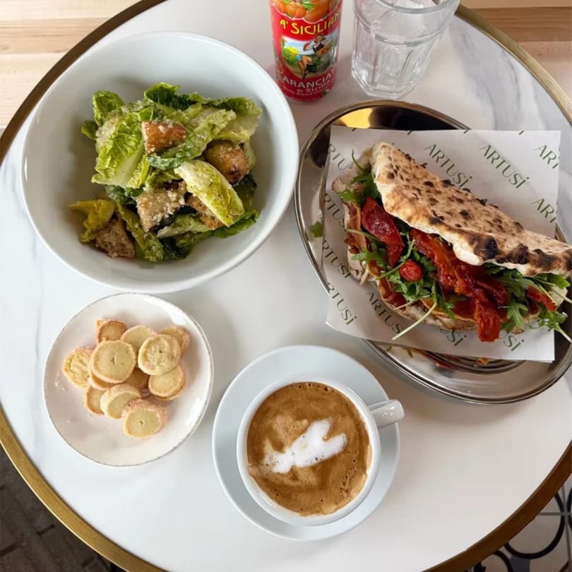 A flatbread sandwich, Caesar salad, cookies, coffee, and orange juice on a white marble table.