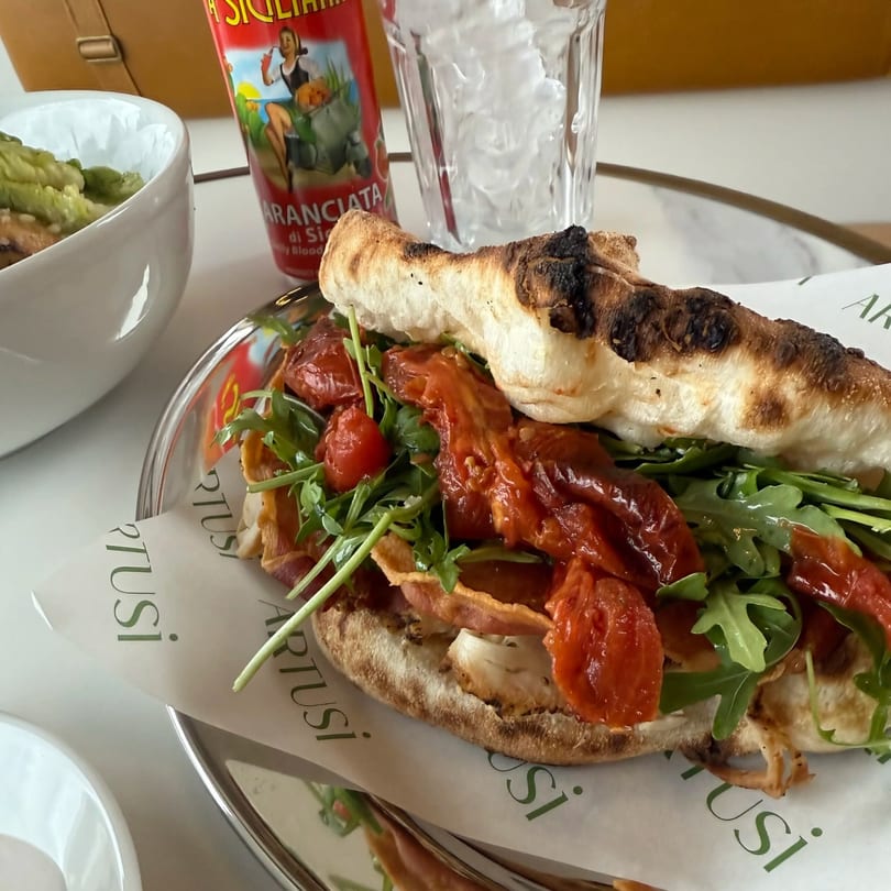 A sandwich with arugula, sun-dried tomatoes, and bacon on focaccia bread, with a drink and side.