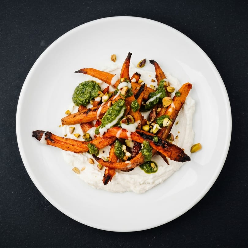 Roasted carrots with green pesto, white sauce, and pistachios on a white plate.